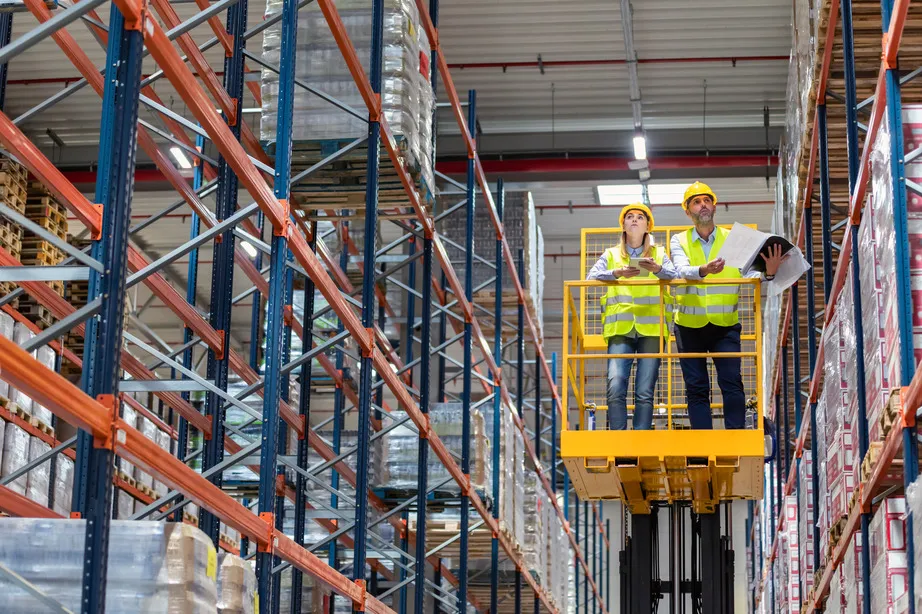 Warehouse inspectors on a lift platform checking inventory on high storage racks.